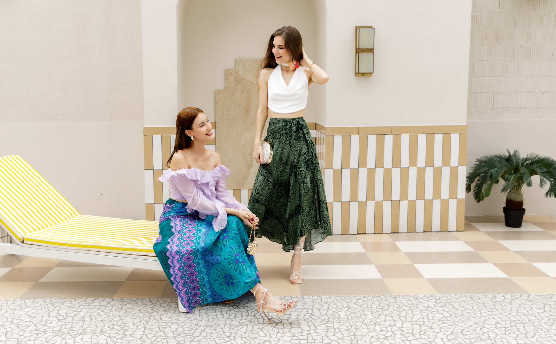 Two models wearing blue and green patterned skirts in a bright courtyard